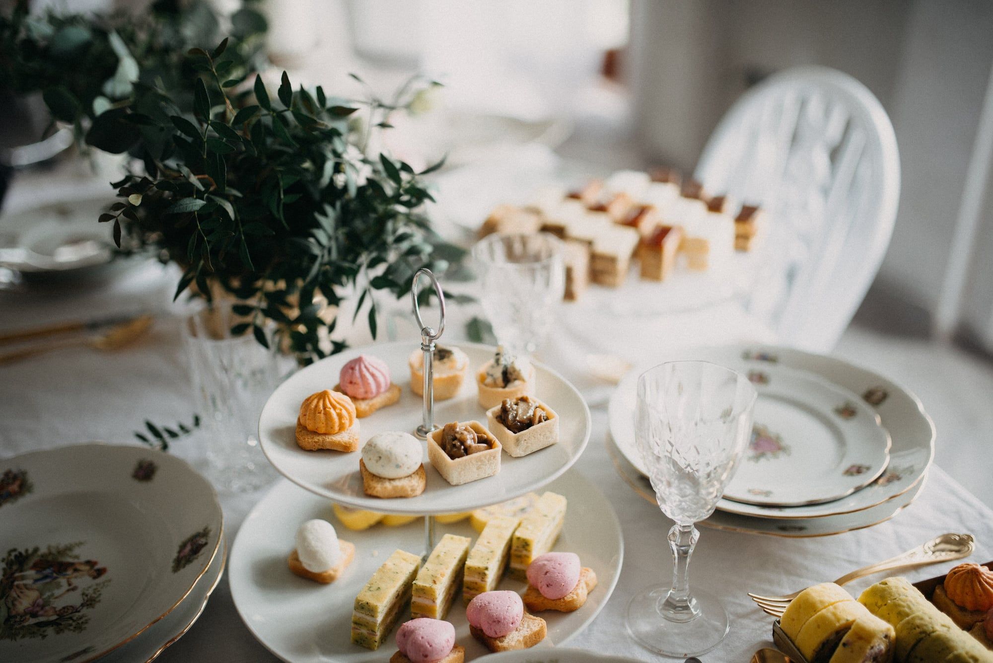 A tiered stand of small cakes and pastries among patterned china and greenery on a celebration table.