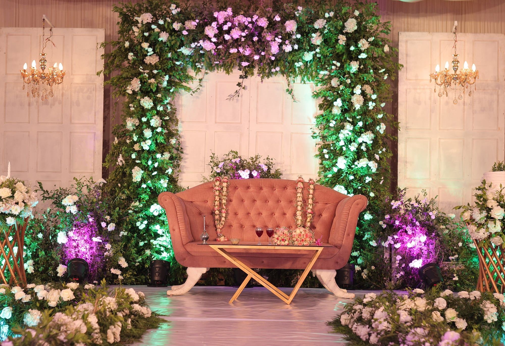 A wedding stage with a coral sofa beneath an arch of roses and greenery, flanked by chandeliers.