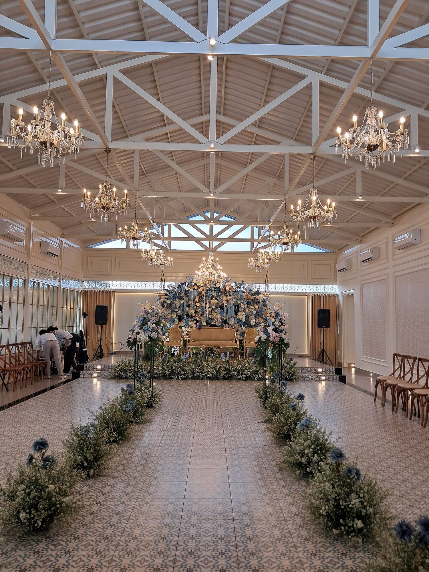 A long aisle lined with low flowers under rows of chandeliers, leading to a floral stage.