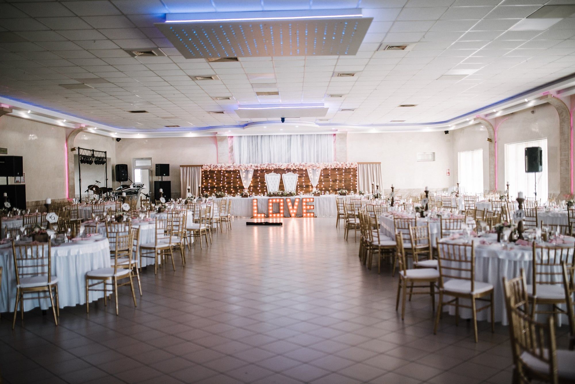 A wide hall set for a wedding, with rows of dressed round tables leading to a decorated head table.