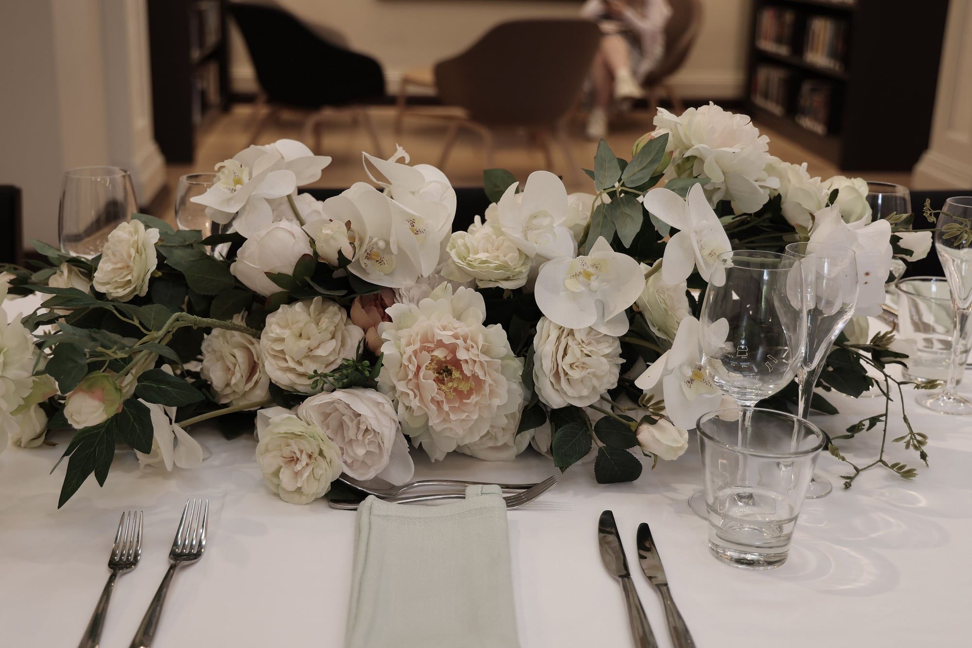 A low centrepiece of white roses and orchids along a table set with silver cutlery and glassware.