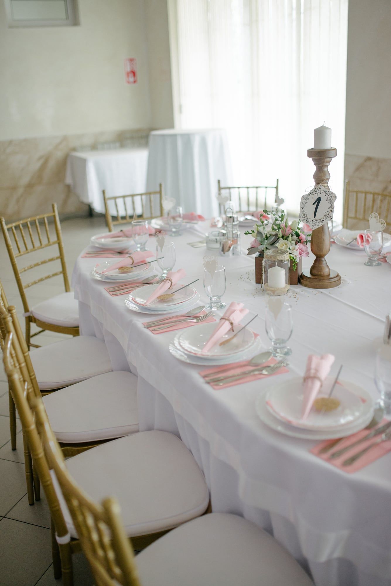 A long table in white linen with blush napkins and gold chairs, beside a bright window.