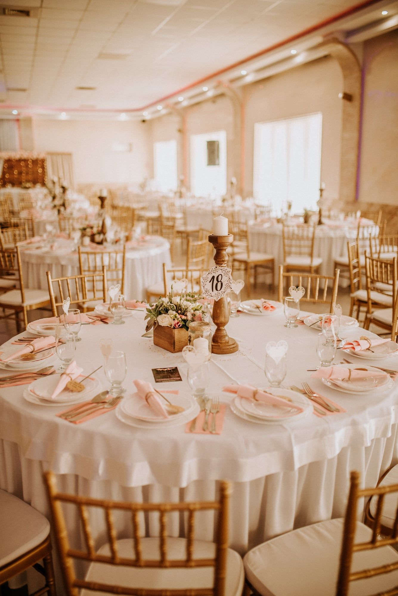 Round banquet tables dressed in ivory cloth with gold chairs and small floral centrepieces, in a bright hall.